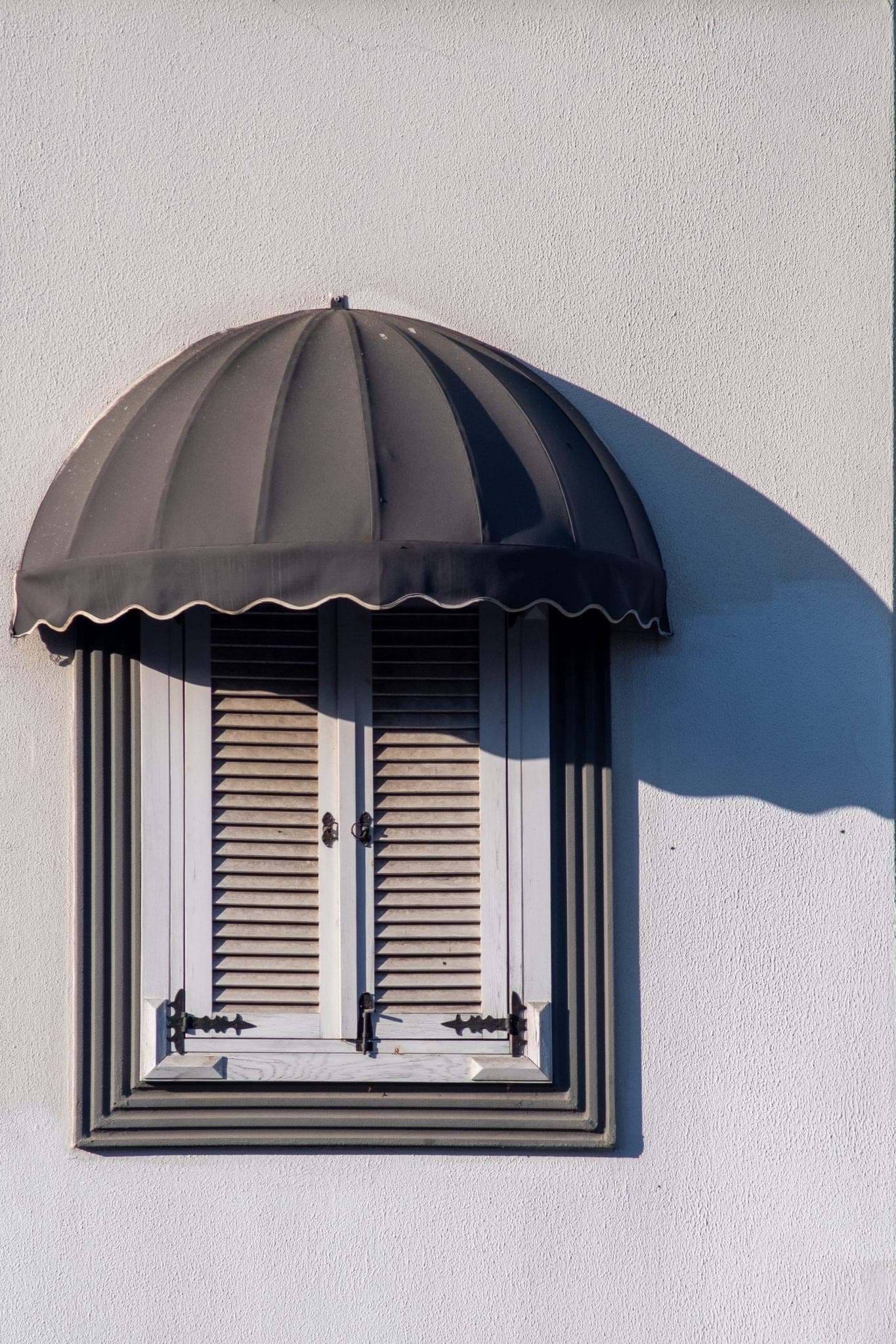 A dark scalloped awning over a shuttered window on a sunlit white wall