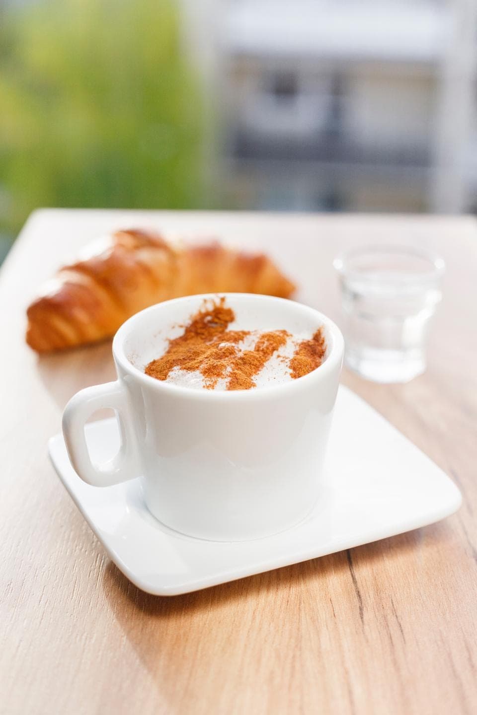 A cappuccino dusted with cinnamon next to a croissant on a white table