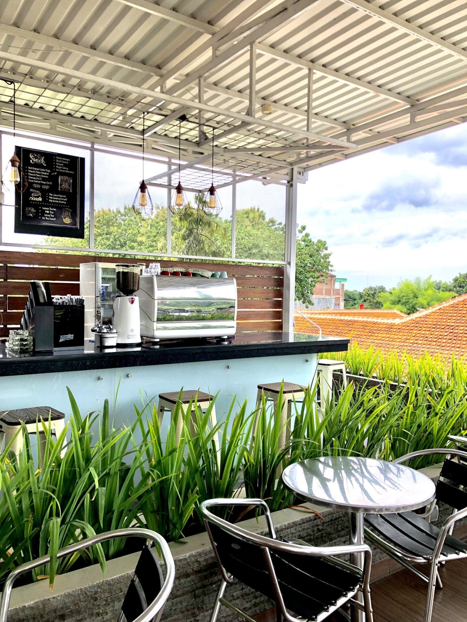A bright rooftop coffee bar with an espresso machine, metal stools and tall grasses in planters