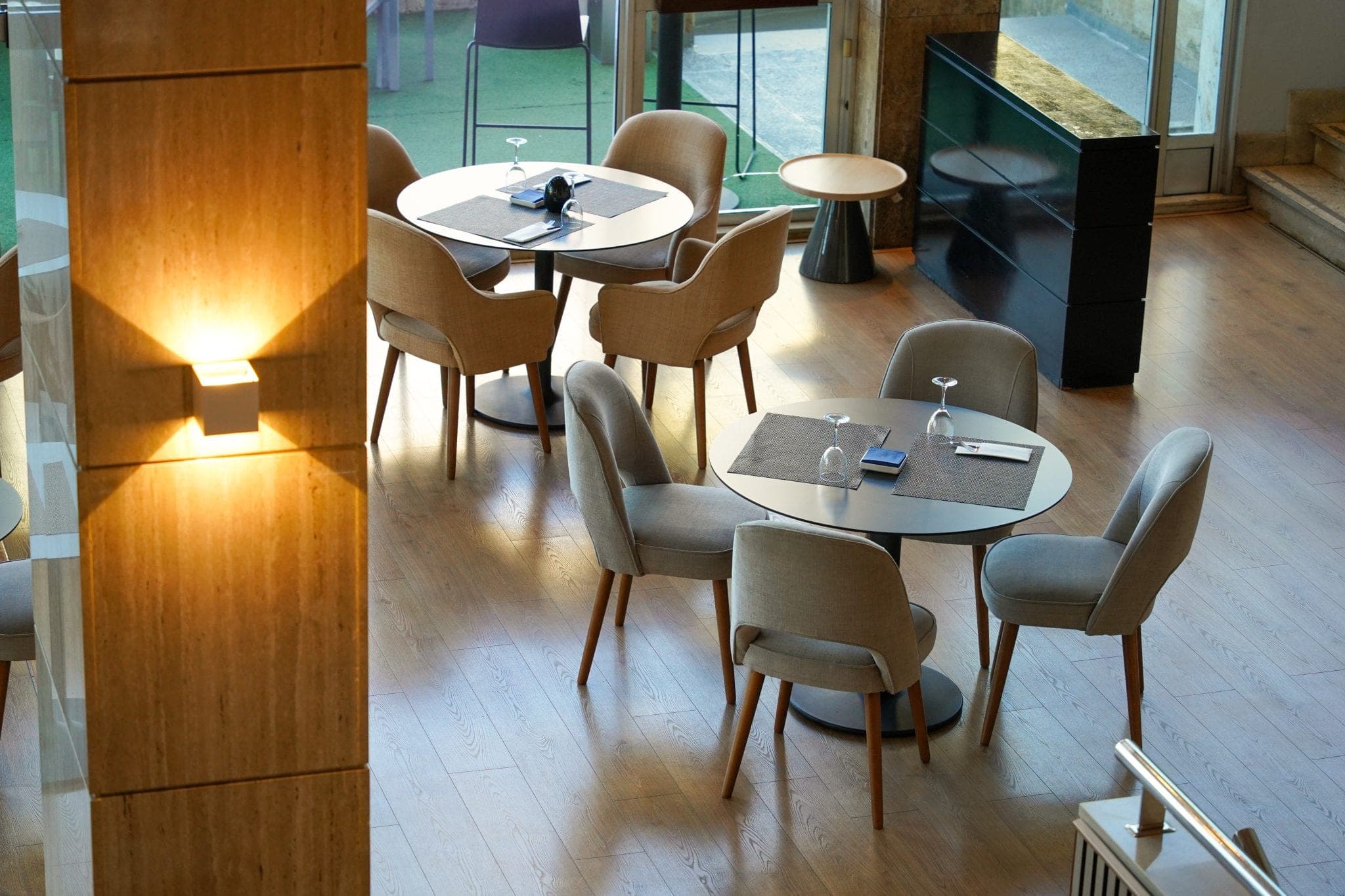 A light-filled café room with round tables, upholstered chairs and a warm wall lamp